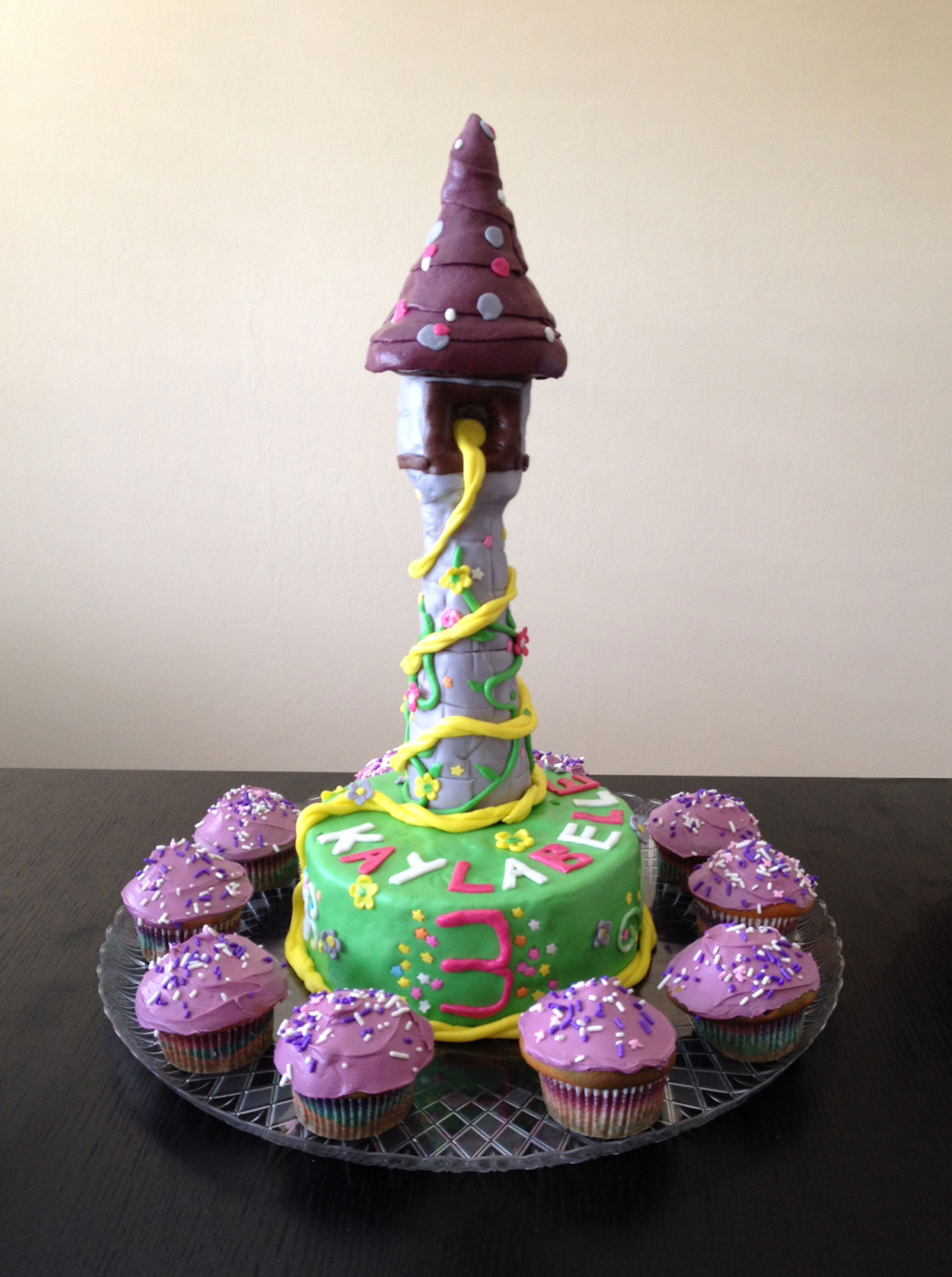 birthday cake shaped into Rapunzel's tower with flowers decoration and long blond hair coming out of the window